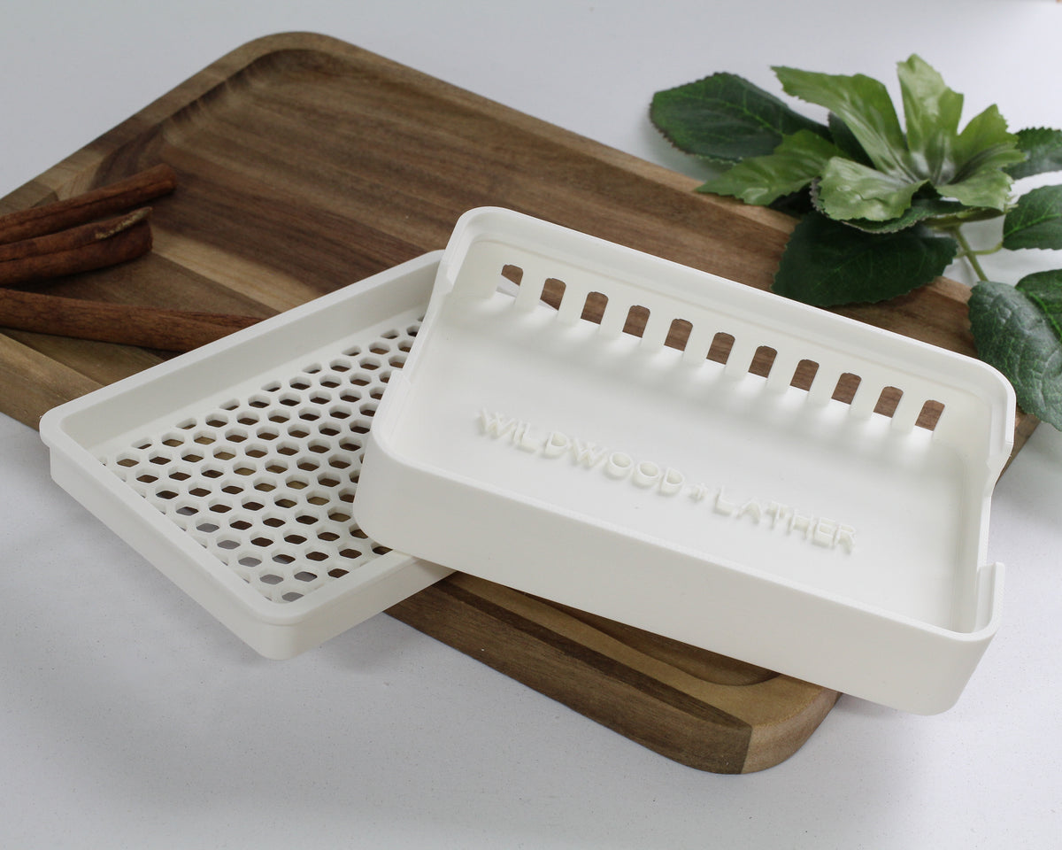 White soap dish with 'Wildwood & Lather' branding on a wooden tray with cinnamon sticks and a plant in the background.